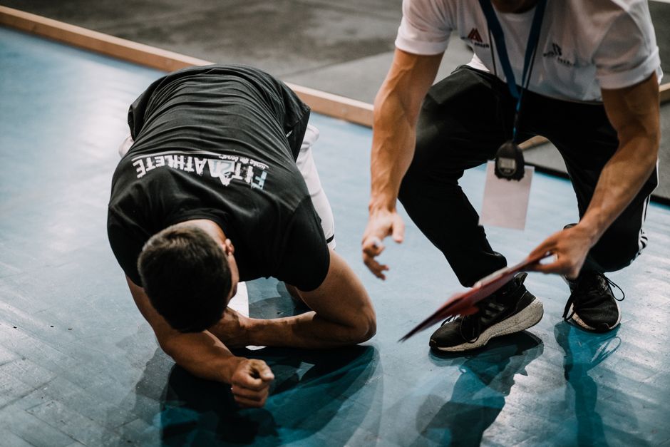 An athlete being assisted by a coach during an indoor training session on a blue floor.